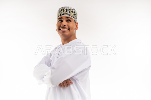 النظر الكاميرا بإيماءات السعادة والسرور, الاهتمام بالأناقة والمظهر الخارجي, تعابير الثقة والاعتزاز بالنفس, بورتريه لرجل عربي خليجي عماني مبتسم يرتدي الدشداشة والكمه يقف باستقامة واعتدال وأيدي متقاطعة, خلفية بيضاء
