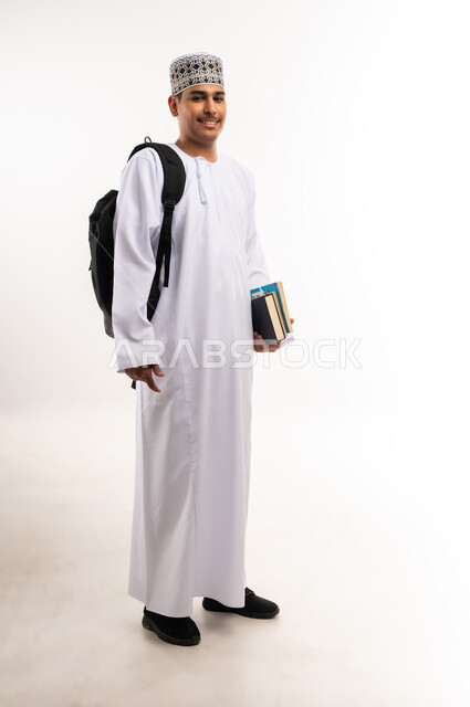 النظر بإيماءات السعادة والسرور, الاستعداد للعام الجامعي الجديد, التعليم العالي في الجامعات والكليات الأكاديمية في سلطنة عمان,  بورتريه لطالب عربي خليجي عماني يرتدي الدشداشة والكمة يحمل حقيبة على ظهره وبيديه الكتب, الجسم بالطول الكامل، خلفية بيضاء