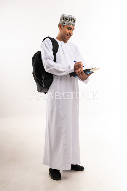 الاستعداد للعام الجامعي الجديد, الجامعات والكليات الأكاديمية في سلطنة عمان, بورتريه لطالب عربي خليجي عماني يرتدي الدشداشة والكمة يحمل حقيبة على ظهره يمسك بيده ورقة وقلم يقوم بمتابعة الدروس اليومية, الجسم بالطول الكامل, خلفية بيضاء