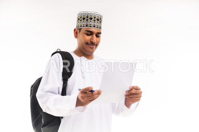 الجامعات والكليات الأكاديمية في سلطنة عمان, الاستعداد للعام الجامعي الجديد,  بورتريه مقرب لطالب عربي خليجي عماني يرتدي الدشداشة والكمة يحمل حقيبة على ظهره يمسك بيده ورقة وقلم يقوم بمتابعة الدروس اليومية, خلفية بيضاء