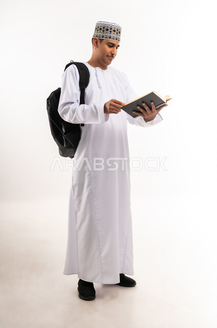 Interest in browsing useful books, love of reading, university education in the Sultanate of Oman, reviewing daily lessons, the concept of diligence and excellence, portrait of an Arab Gulf Omani university student wearing a dishdasha and a kummah, carrying a bag on his back and a book in his hand and studying lectures, white background