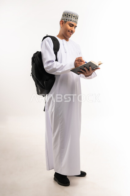 الاهتمام بتصفح الكتب المفيدة، حب المطالعة والقراءة، التعليم الجامعي في سلطنة عمان, مراجعة الدروس اليومية, مفهوم الاجتهاد والتفوق, بورتريه لطالب جامعي عربي خليجي عماني يرتدي الدشداشة والكمة يحمل حقيبة على ظهره وبيده كتاب ويقوم بدراسة المحاضرات, خلفية بيضاء