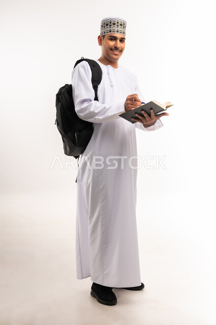 Interest in browsing useful books, love of reading, university education in the Sultanate of Oman, reviewing daily lessons, the concept of diligence and excellence, portrait of an Arab Gulf Omani university student wearing a dishdasha and a kummah, carrying a bag on his back and a book in his hand and studying lectures, white background