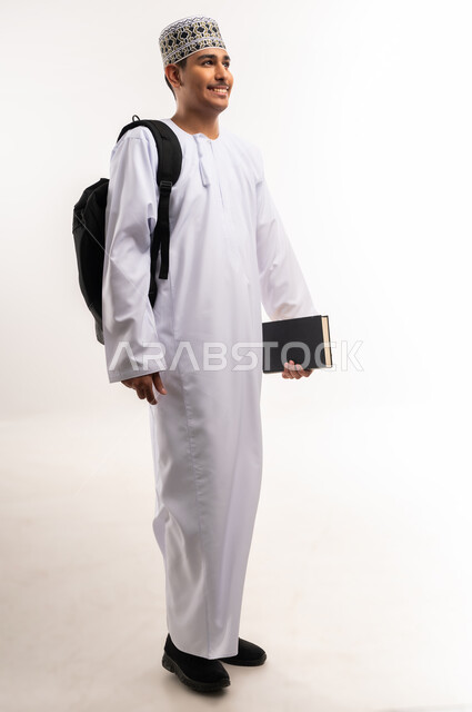 النظر بإيماءات السعادة والسرور, الاستعداد للعام الجامعي الجديد, التعليم العالي في الجامعات والكليات الأكاديمية في سلطنة عمان,  بورتريه لطالب عربي خليجي عماني يرتدي الدشداشة والكمة يحمل حقيبة على ظهره وبيديه الكتب, الجسم بالطول الكامل، خلفية بيضاء