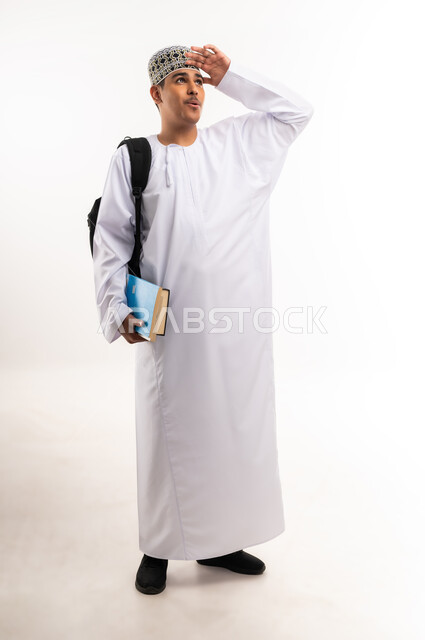النظر لمكان بعيد, تعابير محاولة رؤية شيء ما, مفهوم التطلع للمستقبل, التعليم الجامعي في الإمارات, بورتريه لطالب عربي خليجي عماني يرتدي الدشداشة والكمة يحمل حقيبة على ظهره والكتب الجامعية بيده, الجسم بالطول الكامل، خلفية بيضاء