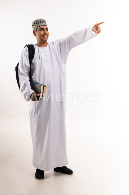 الإشارة بإصبع السبابة إلى شيء ما, الاستعداد للعام الجامعي الجديد, التعليم الاكاديمي في سلطنة عمان, بورتريه لطالب جامعي عربي خليجي عماني يرتدي الدشداشة والكمة ينظر إلى الأعلى ويحمل حقيبة على ظهره والكتب بيديه, الجسم بالطول الكامل, خلفية بيضاء