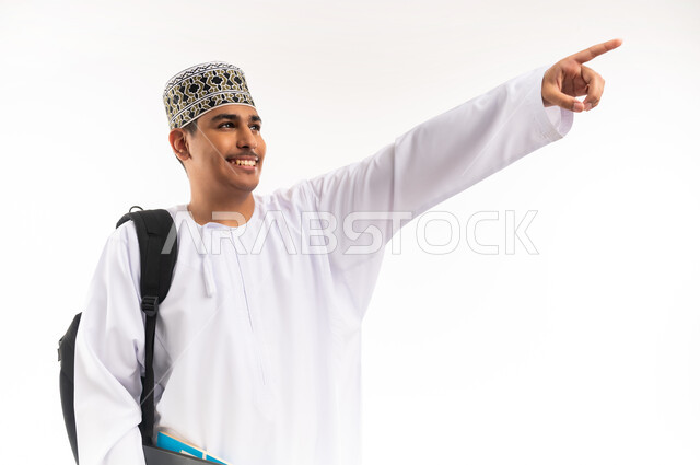 الاستعداد للعام الجامعي الجديد, الإشارة بإصبع السبابة إلى شيء ما, التعليم الاكاديمي في سلطنة عمان, بورتريه مقرب لطالب جامعي عربي خليجي عماني يرتدي الدشداشة والكمة ينظر إلى الأعلى ويحمل حقيبة على ظهره والكتب بيديه, خلفية بيضاء
