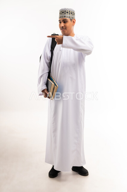 الإشارة بإصبع السبابة إلى شيء ما, الاستعداد للعام الجامعي الجديد, التعليم الاكاديمي في سلطنة عمان, بورتريه لطالب جامعي عربي خليجي عماني يرتدي الدشداشة والكمة يحمل حقيبة على ظهره والكتب بيديه, الجسم بالطول الكامل, خلفية بيضاء