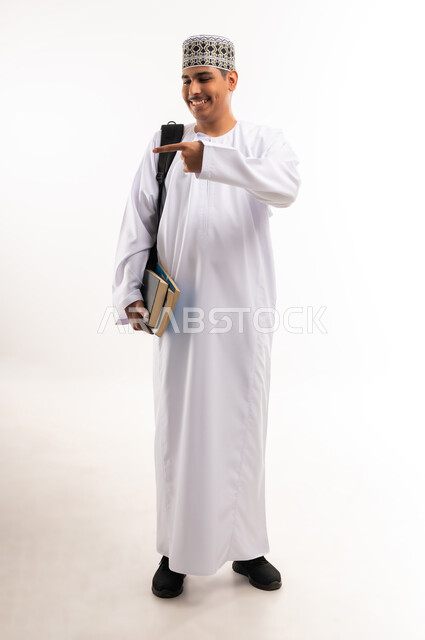 Pointing index finger at something, preparing for the new university year, academic education in the Sultanate of Oman, portrait of an Arab Gulf Omani university student wearing a dishdasha and a turban carrying a bag on his back and books in his hands, full body, white background
