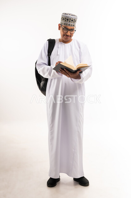 الاهتمام بتصفح الكتب المفيدة، حب المطالعة والقراءة، التعليم الجامعي في سلطنة عمان, مراجعة الدروس اليومية, مفهوم الاجتهاد والتفوق, بورتريه لطالب جامعي عربي خليجي عماني يرتدي الدشداشة والكمة يحمل حقيبة على ظهره وبيده كتاب ويقوم بدراسة المحاضرات, خلفية بيضاء
