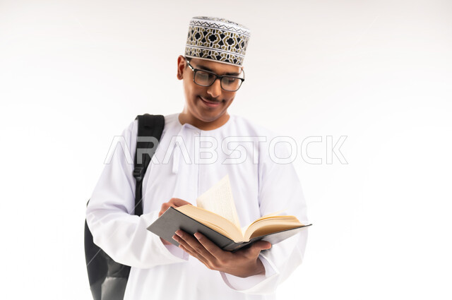 الاهتمام بتصفح الكتب المفيدة، حب المطالعة والقراءة، التعليم الجامعي في سلطنة عمان, مراجعة الدروس اليومية, مفهوم الاجتهاد والتفوق, بورتريه لطالب جامعي عربي خليجي عماني يرتدي الدشداشة والكمة يحمل حقيبة على ظهره وبيده كتاب ويقوم بدراسة المحاضرات, خلفية بيضاء