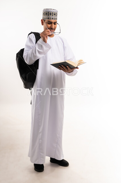 الاهتمام بتصفح الكتب المفيدة، حب المطالعة والقراءة، التعليم الجامعي في سلطنة عمان, مراجعة الدروس اليومية, مفهوم الاجتهاد والتفوق, بورتريه لطالب جامعي عربي خليجي عماني يرتدي الدشداشة والكمة يحمل حقيبة على ظهره وبيده كتاب ويقوم بدراسة المحاضرات, خلفية بيضاء