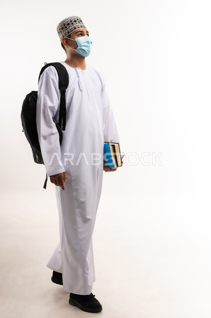 نشر الوعي الصحي, القيام بالإجراءات الوقائية لتجنب الإصابة بفايروس كورونا، التعليم الجامعي في سلطنة عمان، بورتريه لطالب جامعي عربي خليجي عماني يرتدي الدشداشة والكمة مع الكمامة الطبية يحمل حقيبة على ظهره وبيديه الكتب, الجسم بالطول الكامل، خلفية رمادية