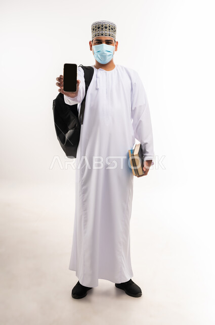 استخدام التكنولوجيا و التقنيات الحديثة المتطورة، بورتريه لشاب عربي خليجي عماني يرتدي الدشداشة و الكمه والكمامة يقوم بعرض شاشة سوداء فارغة عبر الهاتف المحمول،النظر الى الكاميرا بتعابير السرور، صورة الجسم بالطول الكامل, خلفية بيضاء