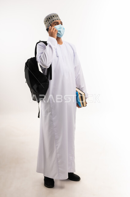 الدردشة والتواصل مع الاصدقاء، استخدام الأجهزة والتقنيات الحديثة، بورتريه لشاب عربي خليجي عماني يرتدي الدشداشة و الكمه والكمامة يحمل الكتب ويمسك بيده الهاتف المحمول يقوم بإجراء المكالمات الهاتفية،  صورة الجسم بالطول الكامل، خلفية بيضاء