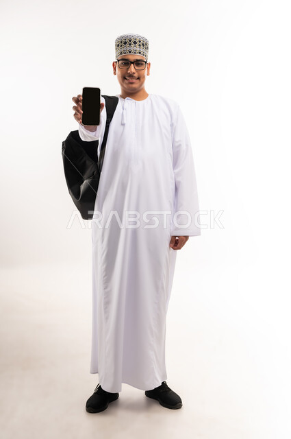 تعابير السعادة والفرح، استخدام الأجهزة والتقنيات الحديثة، بورتريه مقرب لشاب عربي خليجي عماني يرتدي الدشداشة و الكمه يمسك بيده الهاتف المحمول يقوم بعرض شاشة سوداء فارغة بإيماءات السرور، صورة الجسم بالطول الكامل, خلفية بيضاء