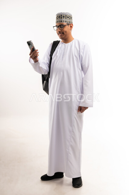 الاستفادة من الأجهزة والتكنولوجيا الحديثة في الاطمئنان على الأقارب، التواصل مع الأهل والأصدقاء وسماع أخبارهم، بورتريه لرجل  عربي خليجي عماني يرتدي الدشداشة والكمه ويمسك الهاتف المحمول بإيماءات السعادة، الجسم بالطول الكامل، خلفية بيضاء