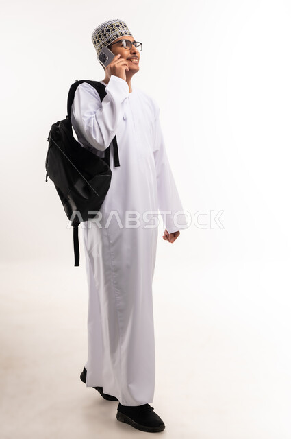استخدام الأجهزة والتقنيات الحديثة، بورتريه لشاب عربي خليجي عماني يرتدي الدشداشة و الكمه يمسك بيده الهاتف المحمول يقوم بإجراء المكالمات الهاتفية، الدردشة والتواصل مع الاصدقاء بإيماءات السعادة والفرح، صورة الجسم بالطول الكامل، خلفية بيضاء