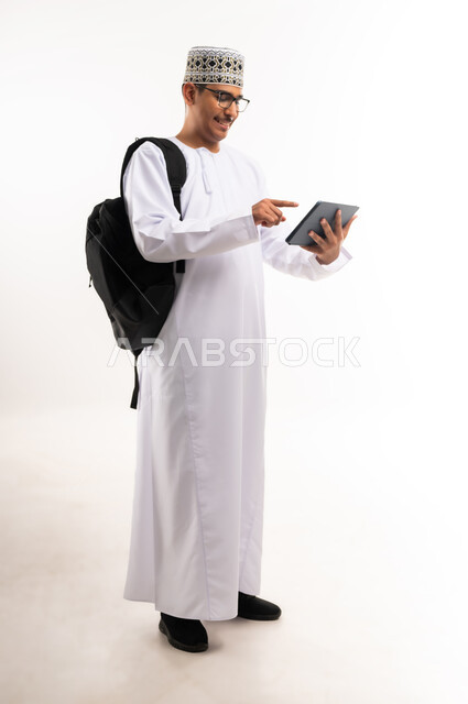 هواية المطالعة والاستكشاف وقضاء وقت الفراغ بالتعلم والثقافة، مفهوم التثقيف والبحث العلمي، بورتريه لرجل عربي خليجي عماني مبتسم يرتدي الدشداشه والكمه يحمل بيده جهاز التابلت المحمول ويقوم بالعمل عليه، صورة الجسم بالطول الكامل, خلفية بيضاء