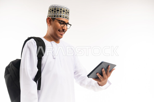 قضاء وقت الفراغ بالتعلم والثقافة، هواية المطالعة والاستكشاف ، مفهوم التثقيف والبحث العلمي، بورتريه مقرب لرجل عربي خليجي عماني مبتسم يرتدي الدشداشه والكمه يحمل بيده جهاز التابلت المحمول ويقوم بالعمل عليه ، خلفية بيضاء