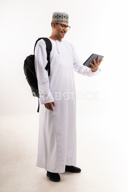 Browsing social media, watching interesting movies and series, chatting with family and friends, using technology and modern techniques, portrait of an Arab Gulf Omani man wearing a dishdasha, a turban and a leather bag holding a tablet in his hand, full body length, white background