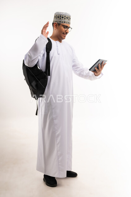 قضاء وقت الفراغ بالتعلم والثقافة، هواية المطالعة والاستكشاف،  مفهوم التثقيف والبحث العلمي، بورتريه لرجل عربي خليجي عماني مبتسم يرتدي الدشداشه والكمه يحمل بيده جهاز التابلت المحمول ويقوم بالعمل عليه، صورة الجسم بالطول الكامل, خلفية بيضاء