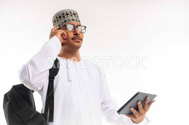 مفهوم التثقيف والبحث العلمي، قضاء وقت الفراغ بالتعلم والثقافة، هواية المطالعة والاستكشاف ، بورتريه مقرب لرجل عربي خليجي عماني مبتسم يرتدي الدشداشه والكمه يحمل بيده جهاز التابلت المحمول ويقوم بالعمل عليه ، خلفية بيضاء