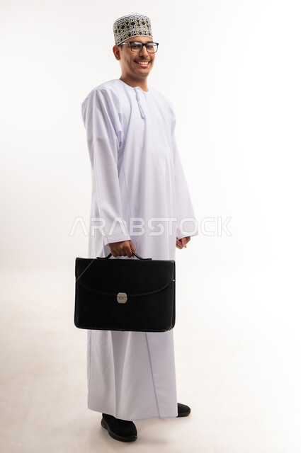 Caring about elegance and appearance, going to work actively, feeling happy to achieve company goals, side portrait of a smiling Omani Gulf Arab businessman wearing a traditional thobe and sleeve holding a handbag, full body image, white background