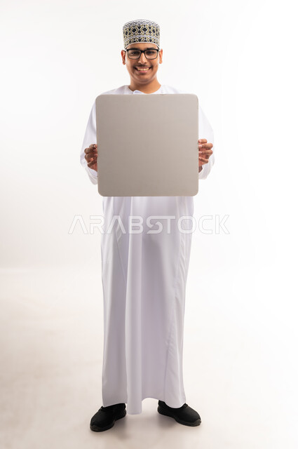 Advertisement and marketing offers, white square mockup, using illustrations and educational tools, portrait of a smiling young Arab Gulf Omani man wearing a dishdasha and a sleeve, holding a blank white square board in his hand and looking at the camera with an expression of pleasure, full body image, white background
