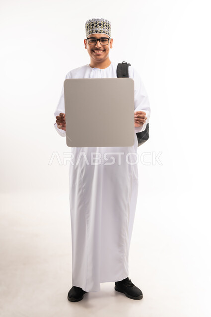 Using illustrations and educational tools, advertising and marketing offers, white square mockup, portrait of a smiling young Arab Gulf Omani man wearing a dishdasha and a sleeve, holding a blank white square board in his hand and looking at the camera with an expression of pleasure, full-length body image, white background