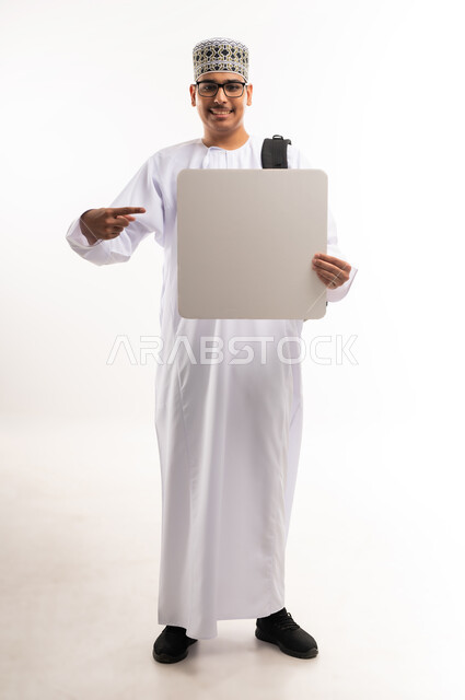 استخدام لوحات توضيحية ووسائل تعليمية, اعلان و عروض تسويقية, موك اب مربع ابيض, بورتريه لشاب مبتسم عربي خليجي عماني يرتدي الدشداشة والكمه يحمل بيده لوحه بيضاء مربعة فارغة و ينظر الى الكاميرا بتعابير السرور, صورة الجسم بالطول الكامل, خلفية بيضاء
