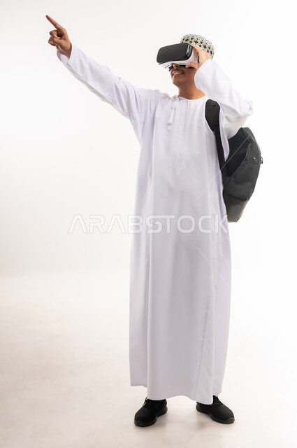 Raising hand with gestures of integration and simulation of the imaginary world, using advanced techniques and technology, portrait of a young Arab Gulf Omani man wearing a dishdasha and an Omani kummah, wearing VR glasses, watching virtual reality movies, spending leisure time in a fun way, full body, white background