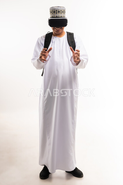 التفاعل والاندماج ومحاكاة العالم الخيالي, بورتريه لشاب عربي خليجي عماني يرتدي الدشداشه والكمه ونظارة الواقع الافتراضي يضع حقيبة الظهر المدرسية, مشاهدة الافلام ثلاثية الابعاد, قضاء أوقات الفراغ بالأنشطة الالكترونية المسلية, الجسم بالطول الكامل, خلفية بيضاء