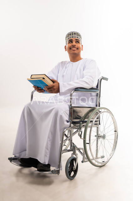اهتمام المجتمع العماني بتعليم ذوي الاحتياجات الخاصة, دمج المعاقين في الجامعات في عمان, بورتريه لشاب عربي خليجي عماني يرتدي الدشداشه والكمه يجلس على الكرسي المتحرك يحمل بيده الكتب الدراسية, النظر إلى شيء ما بسرور, تنمية القدرات العقلية لذوي الهمم, خلفية بيضاء