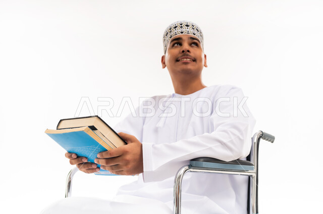 دمج المعاقين في الجامعات في عمان, بورتريه مقرب لشاب عربي خليجي عماني يرتدي الدشداشه والكمه يجلس على الكرسي المتحرك يحمل بيده الكتب الدراسية, النظر إلى شيء ما, تنمية القدرات العقلية لذوي الهمم, اهتمام المجتمع العماني بتعليم ذوي الاحتياجات الخاصة, خلفية بيضاء