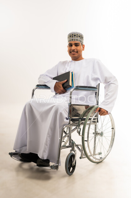 تنمية القدرات العقلية لذوي الهمم, اهتمام المجتمع العماني بتعليم ذوي الاحتياجات الخاصة, دمج المعاقين في الجامعات في سلطنة عمان, بورتريه لشاب عربي خليجي عماني يرتدي الدشداشه والكمه يجلس على الكرسي المتحرك يحمل بيده الكتب الدراسية, خلفية بيضاء