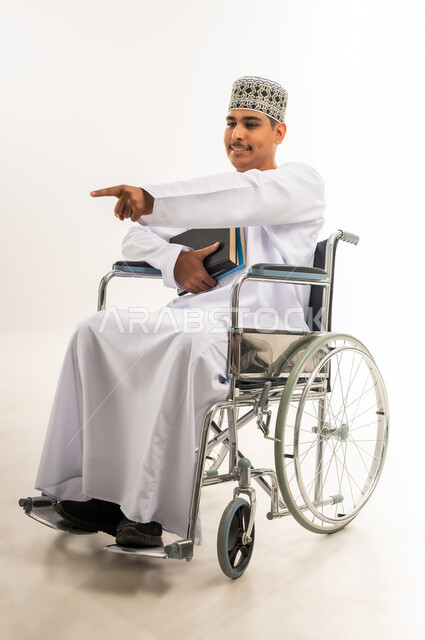 Pointing to something, developing the mental abilities of people with disabilities, Omani society's interest in educating people with special needs, integrating the disabled into universities in the Sultanate of Oman, portrait of a young Gulf Arab Omani man wearing a dishdasha and a cloak sitting in a wheelchair holding school books in his hand, white background