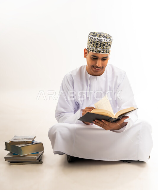 مراجعة الدروس اليومية, مفهوم الاجتهاد والتفوق والنجاح, التعليم الجامعي في سلطنة عمان, بورتريه لطالب جامعي عربي خليجي عماني يرتدي الكمه والدشداشه والنظارة الطبية يجلس على الأرض يحمل بيده كتاب ويقوم بدراسة المحاضرات, خلفية بيضاء