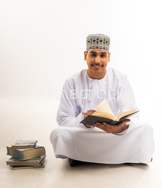 مفهوم الاجتهاد والتفوق والنجاح, التعليم الجامعي في سلطنة عمان, مراجعة الدروس اليومية, بورتريه لطالب جامعي عربي خليجي عماني يرتدي الكمه والدشداشه والنظارة الطبية يجلس على الأرض يحمل بيده كتاب ويقوم بدراسة المحاضرات, النظر إلى الكاميرا بسرور, خلفية بيضاء