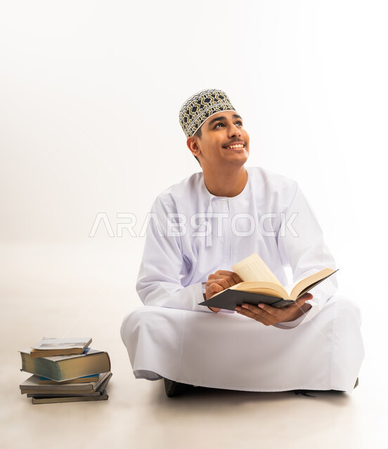 النظر إلى شيء ما بإيماءات السرور, التعليم الجامعي في سلطنة عمان, مفهوم الاجتهاد والتفوق والنجاح, مراجعة الدروس اليومية, بورتريه لطالب جامعي عربي خليجي عماني يرتدي الكمه والدشداشه والنظارة الطبية يجلس على الأرض يحمل بيده الكتاب, خلفية بيضاء