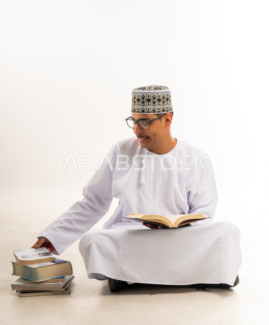 التعليم الجامعي في سلطنة عمان, مفهوم الاجتهاد والتفوق والنجاح, مراجعة الدروس اليومية, بورتريه لطالب جامعي عربي خليجي عماني يرتدي الكمه والدشداشه والنظارة الطبية يجلس على الأرض يحمل بيده كتاب ويقوم بدراسة المحاضرات, خلفية بيضاء