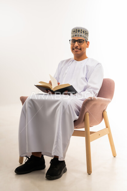 النظر إلى الكاميرا بابتسامة, قضاء أوقات الفراغ بالأنشطة المفيدة, مفهوم التعلم والتفوق والاجتهاد, المناهج التعليمية في سلطنة عمان, الاستمتاع بممارسة هواية المطالعة, بورتريه لشاب عربي خليجي عماني يرتدي الدشداشه والكمه والنظارة يجلس على كرسي مريح, خلفية بيضاء