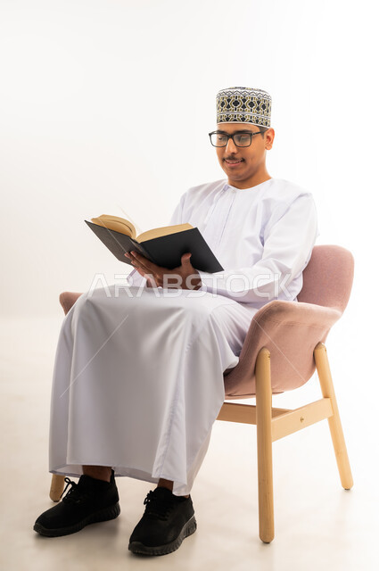 قضاء أوقات الفراغ بالأنشطة المفيدة, مفهوم التعلم والتفوق والاجتهاد, المناهج والمواد التعليمية في سلطنة عمان, الاستمتاع بممارسة هواية المطالعة, بورتريه لشاب عربي خليجي عماني يرتدي الدشداشه والكمه والنظارة الطبية يقوم بقراءة الكتاب باندماج وتركيز, خلفية بيضاء