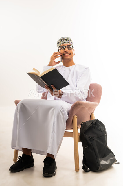 النظر إلى شيء ما واجراء مكالمة صوتية عبر الهاتف المحمول, مراجعة الدروس للمذاكرة والاختبارات المدرسية، تطوير المناهج التعليمية في سلطنة عمان، بورتريه لطالب جامعي عربي خليجي عماني يرتدي الكمه والدشداشة يجلس على كرسي مريح ويمسك بيده الكتاب, خلفية بيضاء
