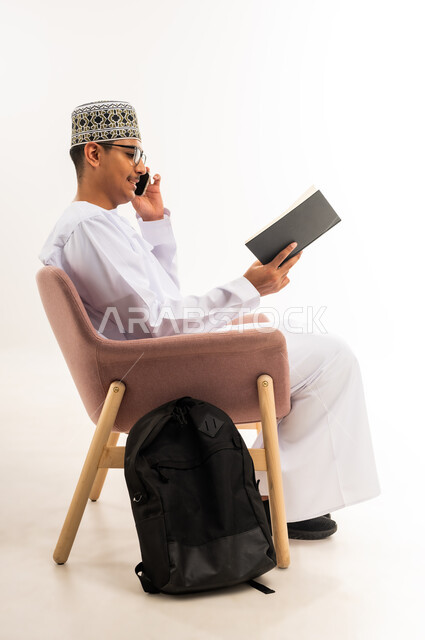 اجراء مكالمة صوتية عبر الهاتف المحمول, مراجعة الدروس للمذاكرة والاختبارات المدرسية، تطوير المناهج التعليمية في سلطنة عمان، بورتريه من الجانب لطالب جامعي عربي خليجي عماني يرتدي الكمه والدشداشة يجلس على كرسي مريح ويمسك بيده الكتاب, خلفية بيضاء
