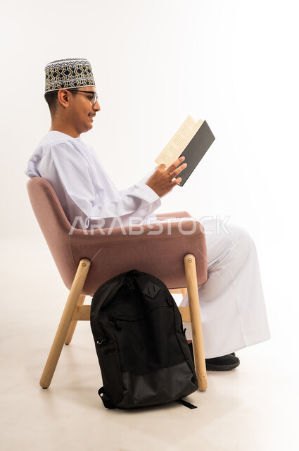 Reviewing lessons for studying and school tests, developing educational curricula in the Sultanate of Oman, expressions of diligence and excellence, side portrait of an Arab Gulf Omani university student wearing a kimmah and a dishdasha sitting on a comfortable chair reading a school book with concentration and integration, white background