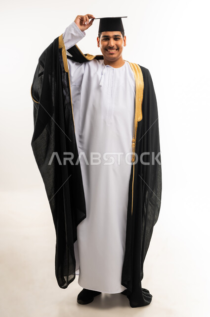 التعليم الاكاديمي في جامعات سلطنة عمان, مفهوم الدراسة والنجاح والتفوق, بورتريه لخريج جامعي عربي خليجي عماني يرتدي العباءة وقبعة التخرج ينظر إلى الكاميرا بابتسامة, مشاعر السرور والسعادة بتحقيق حلم النجاح, صورة الجسم بالطول الكامل, خلفية بيضاء