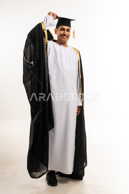مفهوم الدراسة والنجاح والتفوق, بورتريه مقرب لخريج جامعي عربي خليجي عماني يرتدي العباءة وقبعة التخرج ينظر إلى الكاميرا بابتسامة, التعليم الاكاديمي في جامعات سلطنة عمان, مشاعر السرور والسعادة بتحقيق حلم النجاح, صورة الجسم بالطول الكامل, خلفية بيضاء