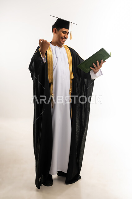 مفهوم الدراسة والنجاح والتفوق, مشاعر السرور والسعادة بتحقيق حلم النجاح, التعليم الاكاديمي في جامعات سلطنة عمان, بورتريه لخريج جامعي عربي خليجي عماني يرتدي العباءة وقبعة التخرج يمسك بيده مشروع التخرج, صورة الجسم بالطول الكامل, خلفية بيضاء