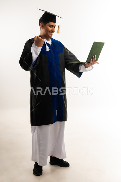 إظهار مشاعر السرور والسعادة بتحقيق حلم النجاح, التعليم الاكاديمي في سلطنة عمان, استخدام الأجهزة والتقنيات الحديثة، بورتريه لخريج جامعي عربي خليجي عماني يرتدي العباءة وقبعة التخرج يقوم باجراء مكالمة فيديو عبر جهاز الحاسوب, صورة الجسم بالكامل, خلفية بيضاء
